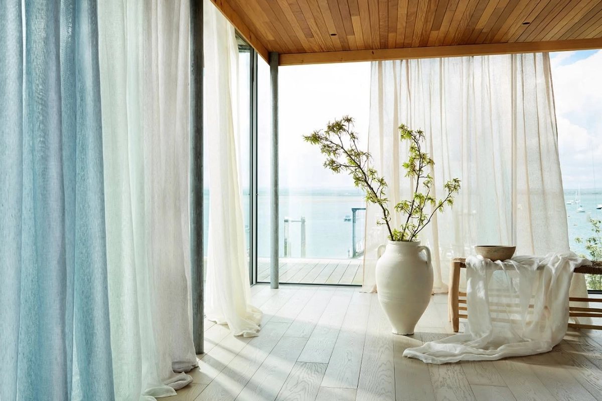Floor-to-ceiling sheer curtains in blue and white framing a bright coastal living space with sea views.