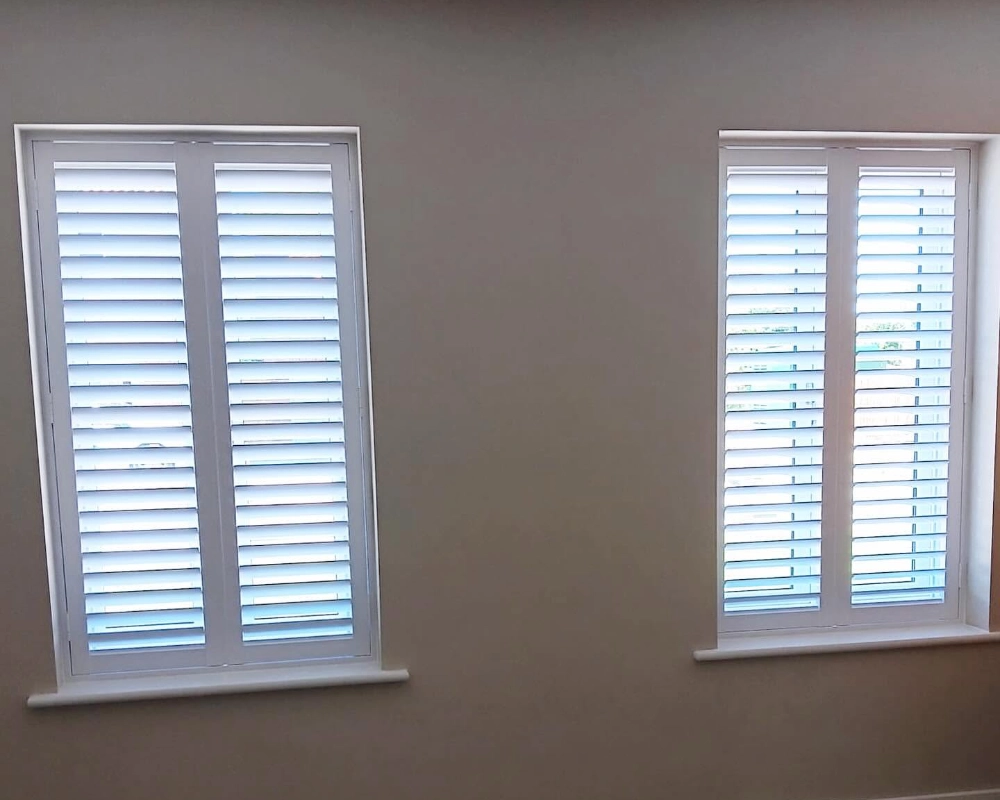 White plantation shutters fitted to two windows in a neutral room with parquet flooring.